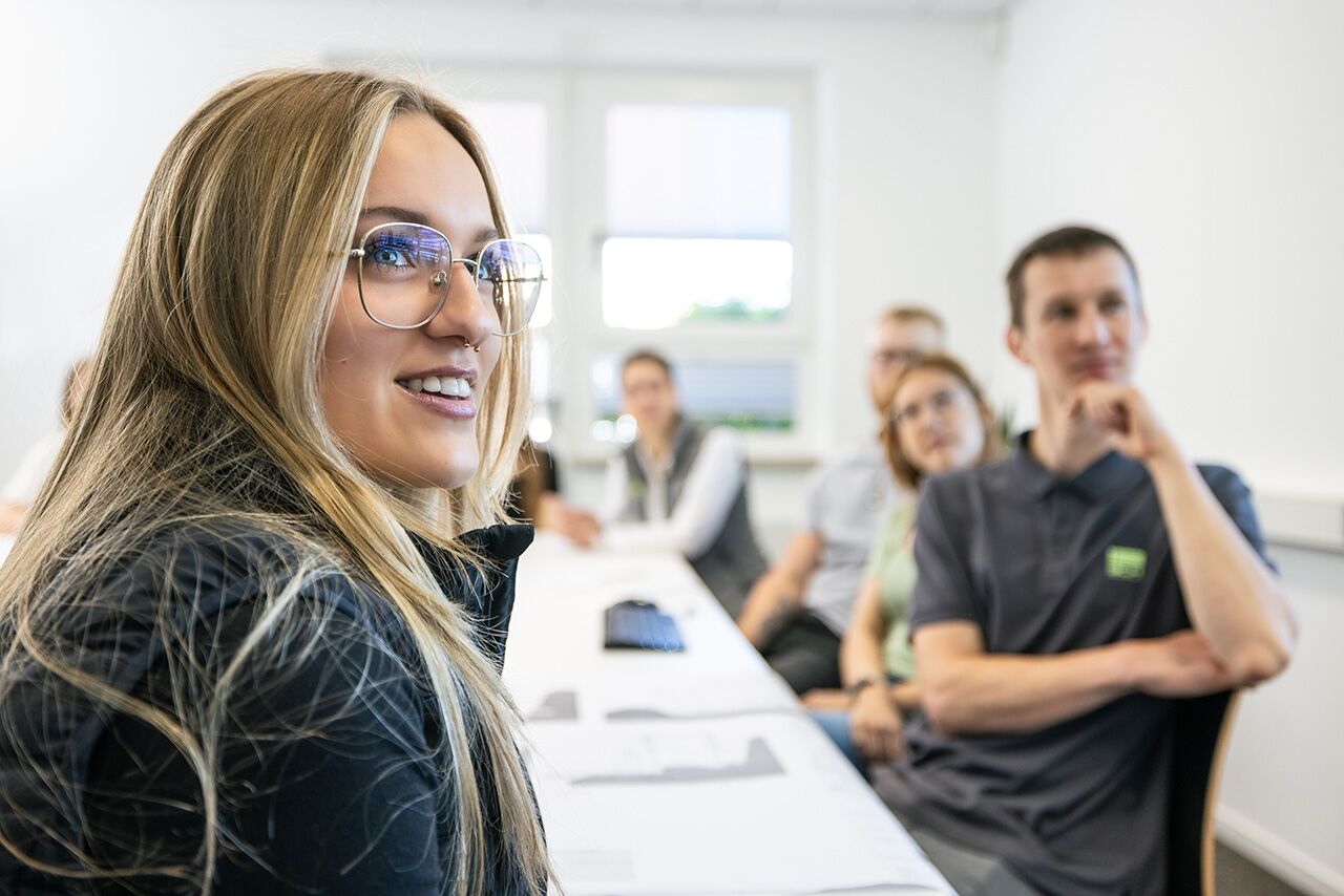Onboarding bei Bau-Consult an langem Meetingtisch, im Vordergrund eine lächelnde blonde Studenten mit Brille