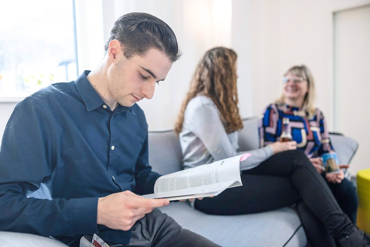 junger Architekt auf grauer Couch liest konzentriert Fachzeitschrift, im Hintergrund zwei Kolleginnen in fröhlicher Unterhaltung