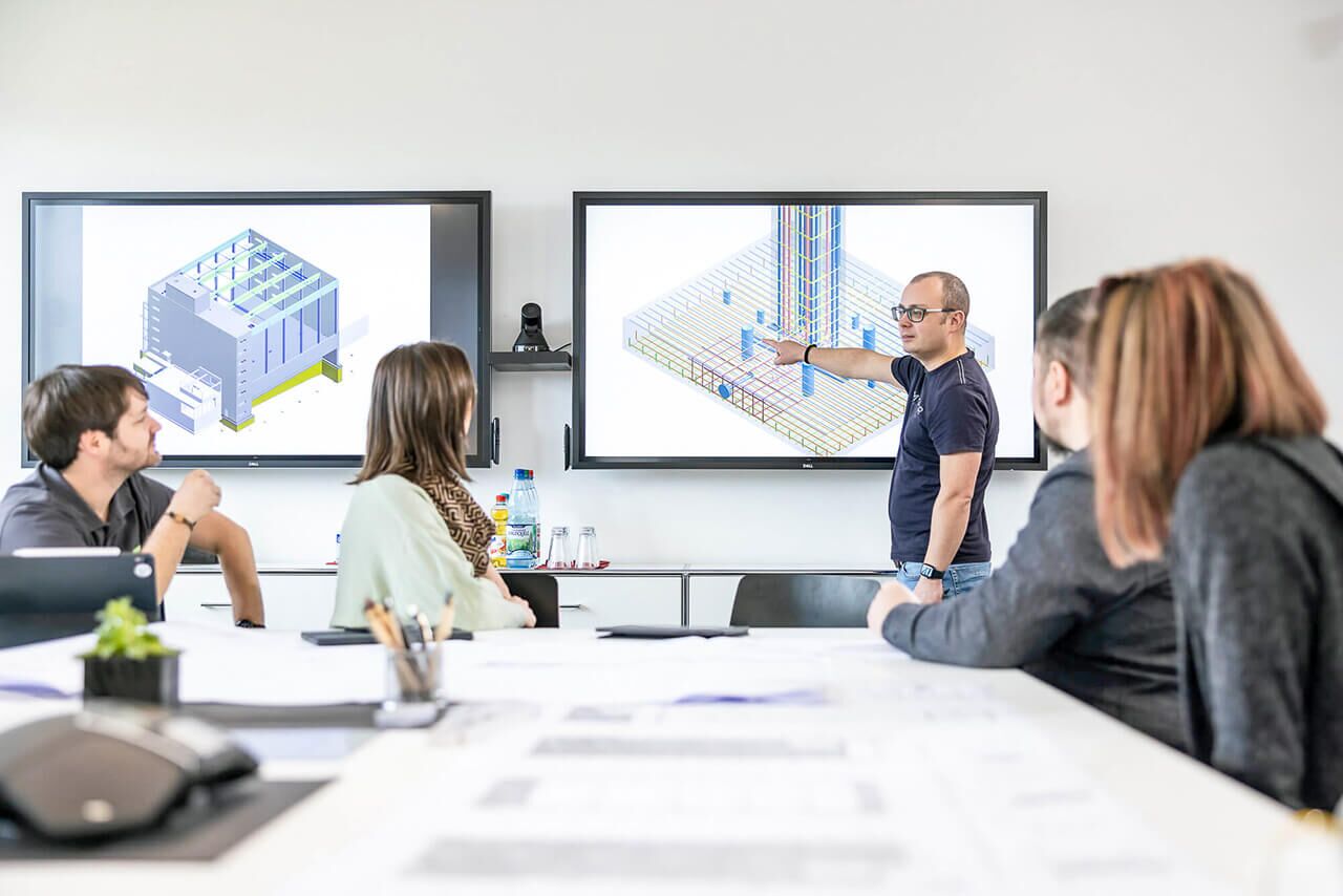 Mitarbeiter in T-Shirt mit schwarzer Brille präsentiert an interaktiven Touch-Displays verschiedene BIM-Modelle, im Vordergrund 4 Personen an großem Tisch